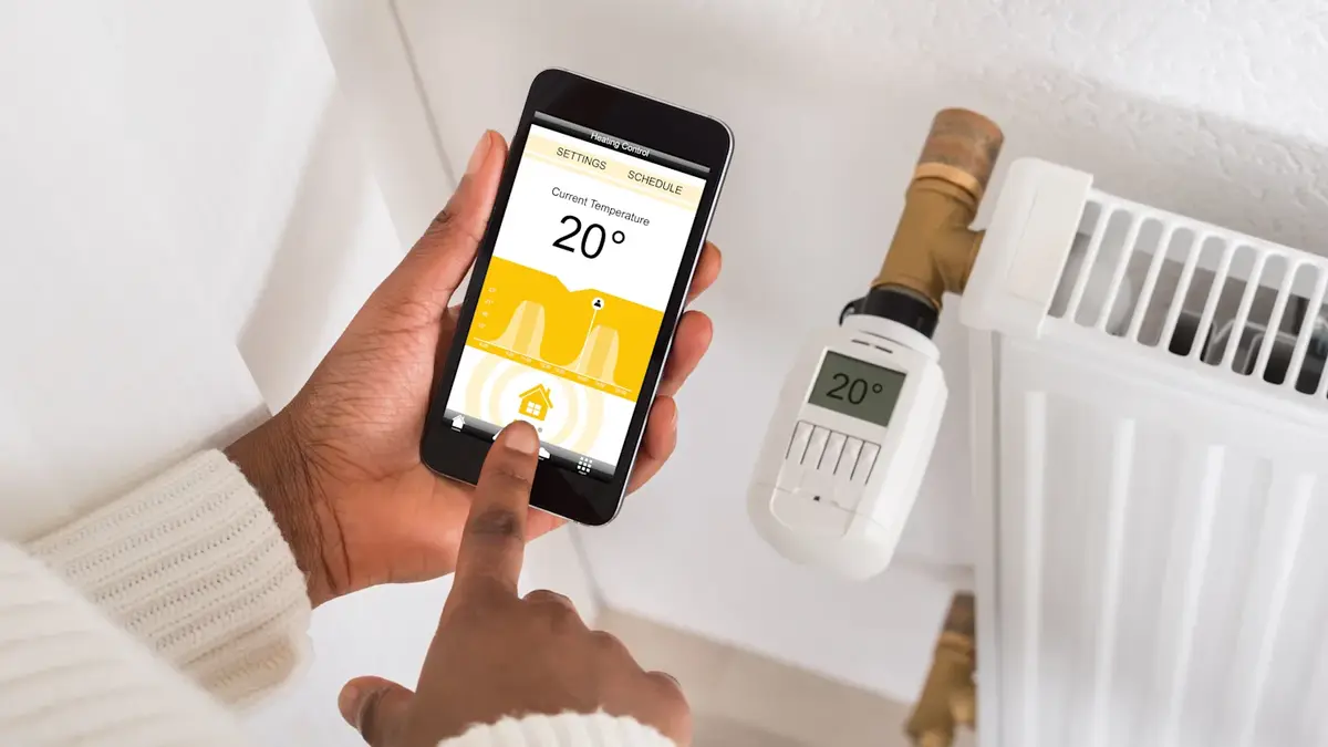 Modernes Smart Thermostat an wei&szlig;er Wohnzimmerwand montiert, Smartphone mit Heizungs-App daneben liegend